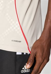 Šviesiai pilka sportinė marškinė su raudonais akcentais ir languota raštą. Turi "AEROREADY" logotipą šalia apvado. Minkšta, drėgmę sugerianti medžiaga.