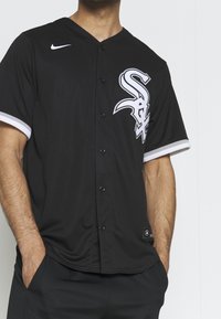 Maillot de baseball noir à manches courtes avec un décolleté en V, présentant des accents blancs, des boutons sur le devant et un logo blanc bien visible sur la poitrine.