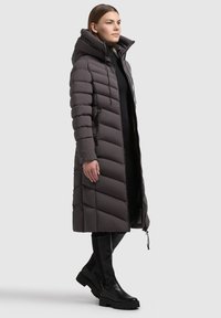 Frau in einem langen, dunkelgrauen, gepolsterten Wintermantel mit Kapuze, schwarzem Kleid und knielangen schwarzen Stiefeln, steht vor einem schlichten Hintergrund.