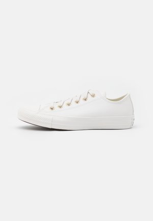 Biały niskoprofilowy sneaker ze złotymi oczkami i białymi sznurowadłami, widziany z boku na jednolitym jasnym tle.