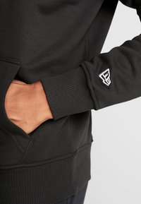 Sudadera negra hecha de una tela suave, con puños y dobladillo acanalados, un bolsillo lateral y un logo blanco en la manga.