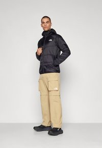 Svart isolerad jacka med huva, beiga cargobyxor med fickor och svarta skor med texturerade sulor.