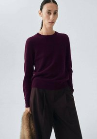 Burgunderfarbener Strickpullover mit Rundhalsausschnitt, getragen mit dunkelbraunen weit geschnittenen Hosen. Die Textur wirkt weich, mit eng anliegenden Ärmeln und einer lockeren Passform.