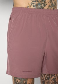 Hellblaue Sportshorts mit elastischem Bund, Seitentaschen und "PROJECT ROCK" Branding nahe dem Saum. Glatte Textur.