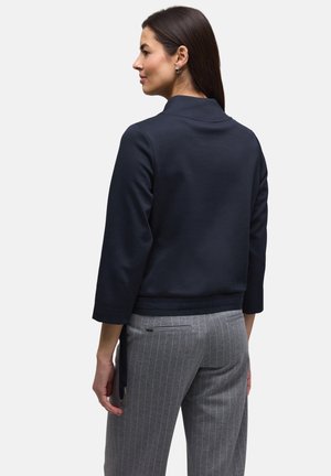 Femme aux cheveux longs et foncés, portant un haut bleu marine à manches longues et un pantalon gris à fines rayures, debout de profil sur un fond neutre.