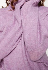 Eine Frau trägt einen weichen, flauschigen lila Pullover, dessen Ärmel ihre Hände bedecken, und ihr lockiges, welliges Haar ist oben sichtbar.