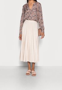 Plisserad krämfärgad midkjol i kombination med en blommig, lätt brun blus. Accessoarer inkluderar en liten rosa handväska och mjuka rosa sandaler.
