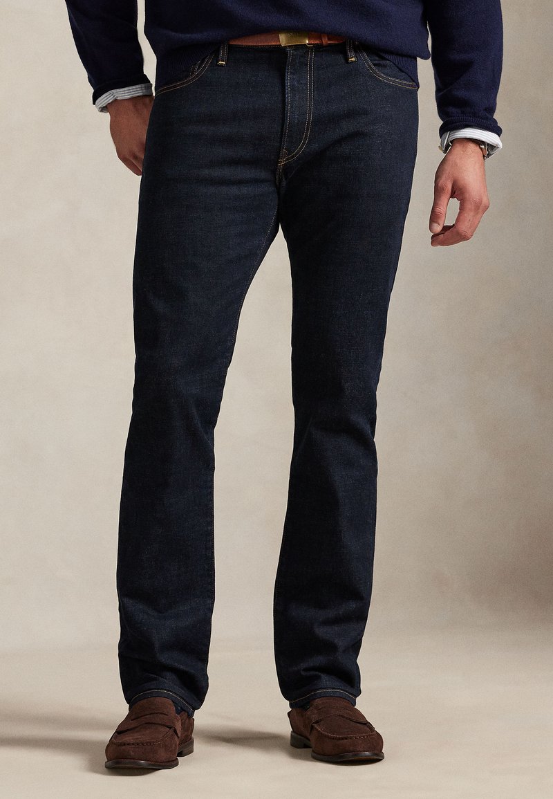 Temno modre kavbojke iz jeansa s ravnim krojem hlačnic, kontrastnim šivanjem, klasičnim slogom s petimi žepi, kombinirane z rjavimi semišnimi čevlji brez vezalk.