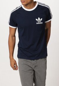 Námořnické tričko s krátkým rukávem vyrobené z bavlny. Disponuje bílým logem Adidas, kontrastním bílým límečkem a pruhovanými rukávy s přiléhavým střihem.