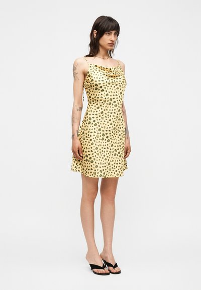 Γυναίκα με σκούρα μαλλιά που φοράει κίτρινο φόρεμα νυχτικό με πράσινες λεοπάρ κηλίδες και μαύρα σανδάλια με τακούνι, στέκεται μπροστά σε μονόχρωμο φόντο.