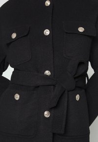 Manteau noir en tissu texturé, avec six boutons décoratifs argentés, deux poches poitrine à rabat et une ceinture à nouer à la taille.