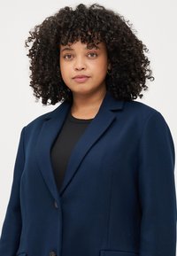 Una giovane donna con capelli ricci indossa un blazer blu navy sopra una maglietta nera, guardando con fiducia verso la macchina fotografica su uno sfondo chiaro.