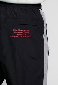 Un pantalon en nylon noir présente un panneau latéral gris. Un texte rouge brodé inclut "Nike International Goddess of Victory" et des détails de localisation.
