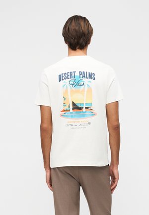 Mand iført hvid t-shirt med farverigt motiv af midten af århundredets villa, palmer og pool, set bagfra mod ensfarvet baggrund.