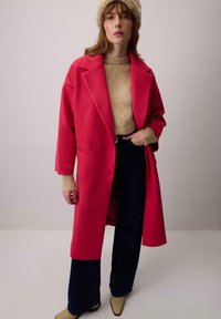 Fukszia gyapjú keverék, kétsoros kabát széles hajtókákkal, oldalsó zsebekkel és laza szabással, bézs kötött pulóver és sötét nadrág felett viselve.