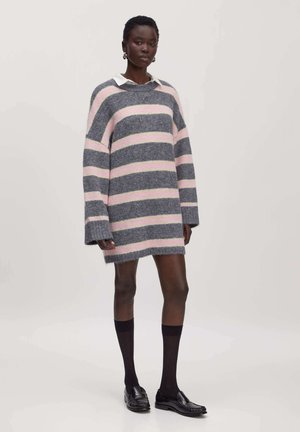 Woman wearing oversized gray and pink striped sweater dress with white collar, black knee-high socks, and black loafers, standing.