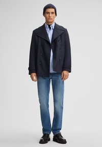 Mann in einem navyfarbenen Peacoat, hellblauem Hemd, blauer Jeans, schwarzen Schuhen und navyfarbenem Beanie, der vor einem schlichten grauen Hintergrund steht.