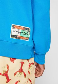 Sweat-shirt bleu avec un patch coloré Tommy Hilfiger et Andy Warhol, associé à un short beige avec un imprimé de homard rouge.