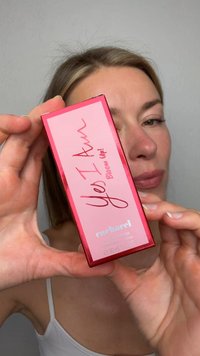 Kvinde holder en lyserød og rød parfumeæske mærket "Yes I Am Bloom Up!" af Cacharel tæt på sit ansigt.