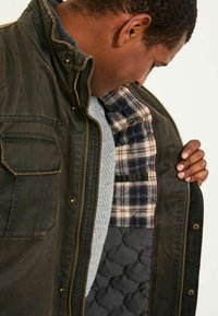 Braune, gesteppten Außenjacke mit Reißverschluss, ausgestattet mit einem platzierten Innenfutter in Blau und Creme. Enthält eine Brusttasche und einen grauen, gerippten Pullover darunter.