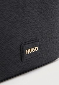 Μαύρη δερμάτινη επιφάνεια με υφή και μια χρυσή πλακέτα λογότυπου «HUGO» ραμμένη στο υλικό.