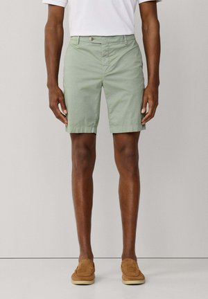 Man draagt lichtgroene knielange shorts, een wit ingestopt T-shirt en lichtbruine instapschoenen, staand tegen een effen achtergrond.