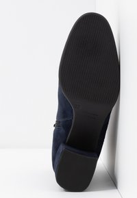 Semelle d'une botte cheville en daim bleu marine montrant une semelle extérieure en caoutchouc noir texturé et un talon bloc, taille 37, positionnée verticalement sur fond blanc.