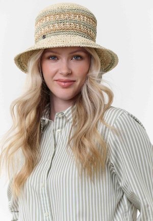 Joven mujer con cabello largo, rubio y ondulado, vestida con una camisa de botones a rayas y un sombrero de sol tejido en beige y verde, sonriendo suavemente.