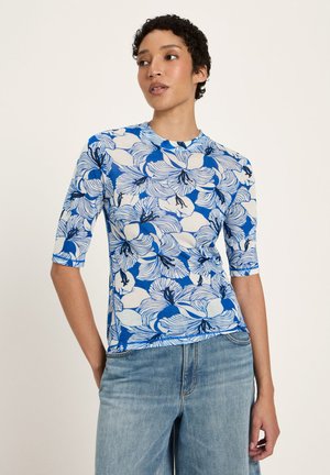 Woman wearing a blue and white floral short-sleeve top paired with light blue denim jeans, standing against a plain background.