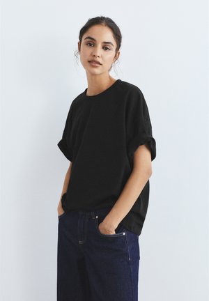 Fiatal, sötét hajú nő laza fekete pólót és sötétkék farmert visel, zsebébe dugott kézzel, egyszínű háttér előtt áll.