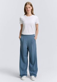 Biele tričko s krátkym rukávom v kombinácii s voľnými, plisovanými modrými nohavicami s hladkou textúrou a bočnými vreckami. Obuv: biele tenisky.