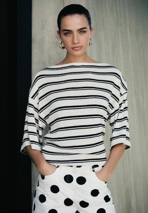 Woman with dark hair in a striped white and black top and white shorts with large black polka dots, standing with hands in pockets against a neutral background.