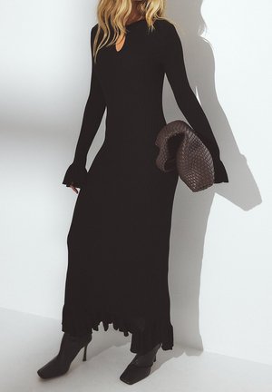 Femme en robe longue noire côtelée et bottes à talons noires tenant une pochette tressée marron foncé sur fond blanc.