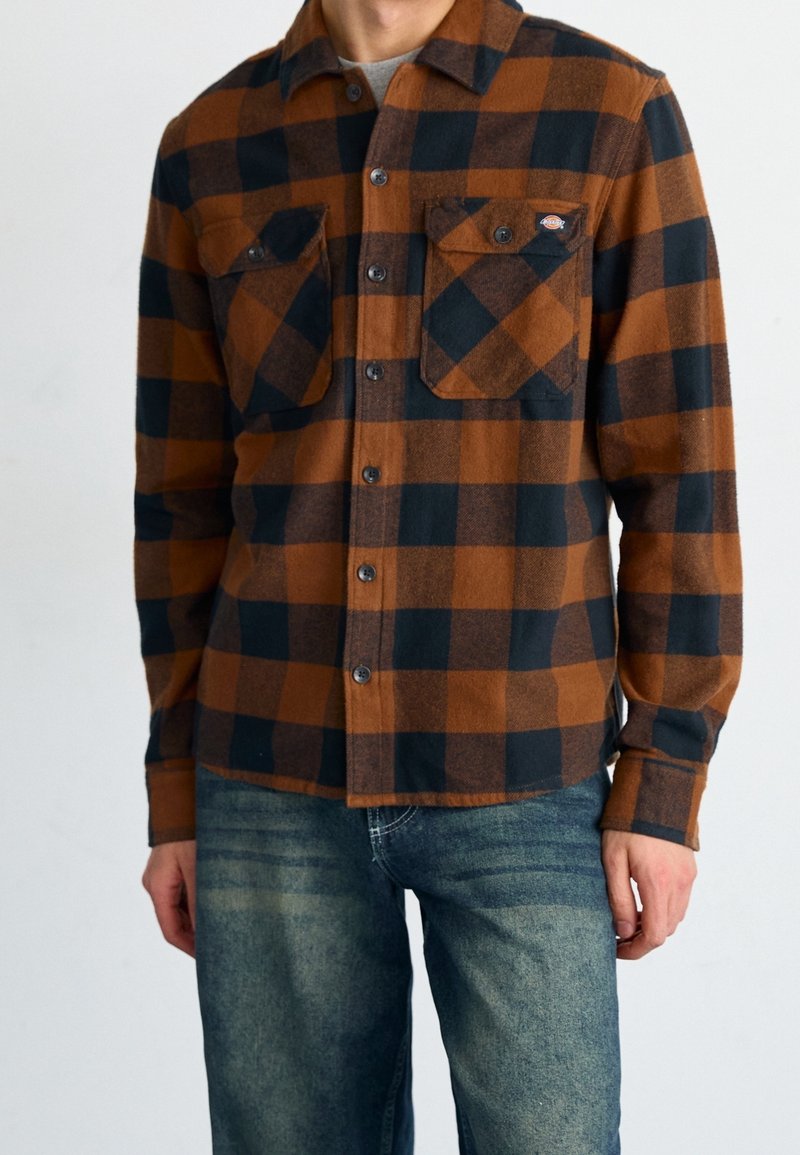 Homme portant une chemise à carreaux marron et noir boutonnée avec deux poches poitrine et un jean bleu sur un fond uni.