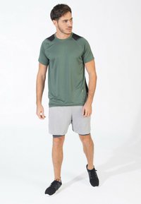 Camiseta atlética verde con acentos negros en los hombros, combinada con pantalones cortos grises. La tela parece ser de secado rápido. Zapatillas deportivas negras completan el atuendo.