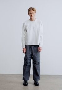 Άσπρο φούτερ με ανάγλυφο λογότυπο, συνδυασμένο με άνετο, σκούρο γκρι, υφασμάτινο παντελόνι. Το μοντέλο φοράει μαύρες χοντρές μπότες. Απλός, μινιμαλιστικός σχεδιασμός.