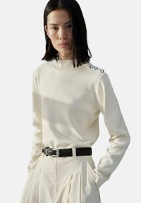 Cream knit sweater with high neck and three decorative buttons on the shoulder. Paired with high-waisted pleated trousers and a black belt.