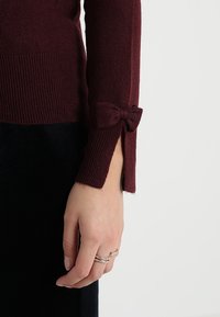 Burgundy sweater with ribbed texture and bow detail on the cuff, featuring a fitted design that extends to the wrist.