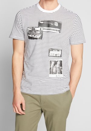 Homme portant un t-shirt blanc rayé de noir avec des motifs musicaux noir et blanc, et un pantalon vert olive.