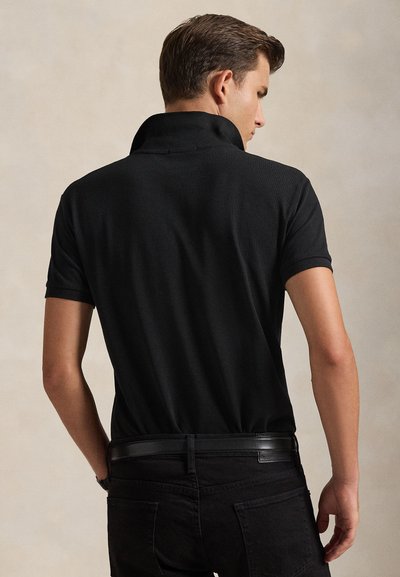 Mann trägt schwarzes kurzärmeliges Poloshirt mit hochgestelltem Kragen und schwarze Hose, von hinten vor neutralem Hintergrund gezeigt.