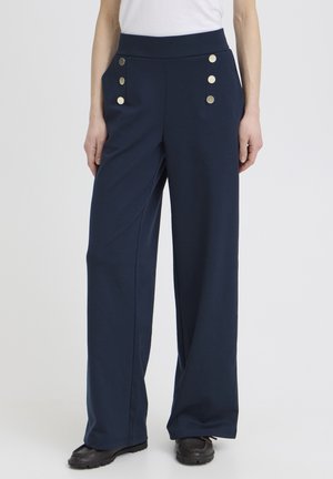 Granatowe spodni szerokie z wysokim stanem i dwoma rzędami po trzy srebrne guziki, noszone z czarnymi butami i białą bluzką.