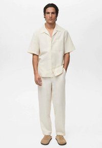 Cămașă scurtă din in bej, cu un buzunar, asortată cu pantaloni ușori în nuanță de bej și pantofi slip-on bej. Stil casual și potrivire confortabilă.