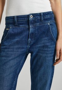 Jeans de mezclilla en azul oscuro con un ligero desgaste, que presentan un diseño clásico de cinco bolsillos, cierre de botón y detalles de costura sutiles.