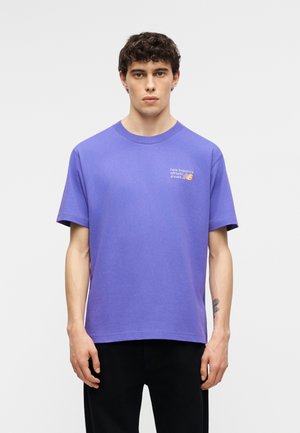 Joven con cabello rizado, vestido con una camiseta deportiva morada de New Balance y pantalones negros, de pie frente a un fondo liso.