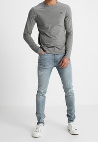 Maglietta grigia a maniche lunghe con scollatura tonda e piccolo logo. Indossata con jeans strappati blu chiaro e sneakers bianche. Stile casual.