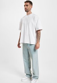 Witte oversized T-shirt van katoen, gecombineerd met lichtblauwe baggy jeans. Model draagt witte sneakers. Eenvoudig, strak ontwerp zonder patronen.