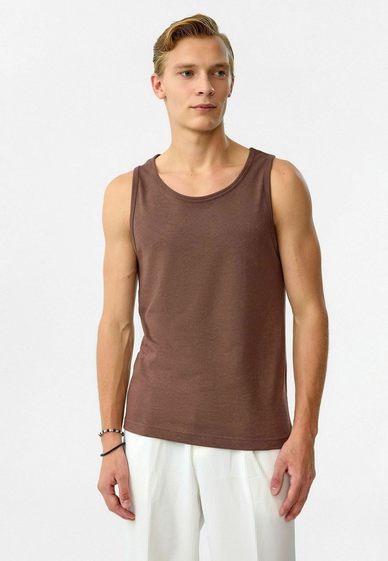 Bruine katoenen tanktop met een ronde hals, slim fit en textuurstof. Draag met witte plissé broek; model staat tegen een neutrale achtergrond.