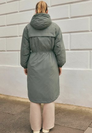 Persoon met blond paardenstaart, gekleed in een lange grijze hoodie, beige wijde broek en witte schoenen, staande op het trottoir naast een witte muur.