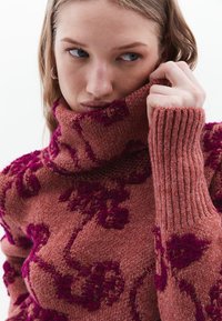 Rosenfarbener Rollkragenpullover aus strukturiertem Strick mit dunkelrosa floralen Akzenten und gerippten Bündchen. Nahaufnahme des Musters und des Stoffs.
