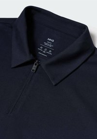 Polo bleu marine avec une fermeture éclair à moitié devant, col pointu et étiquette intérieure indiquant la taille, les consignes d'entretien et la marque MNG.